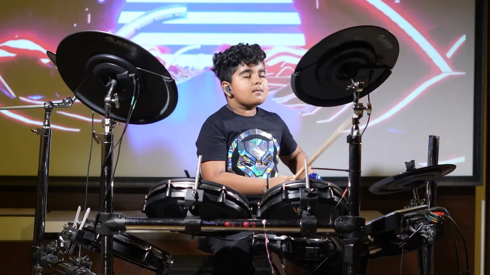 Hridan performing on an electronic drum kit in front of colourful stage lights